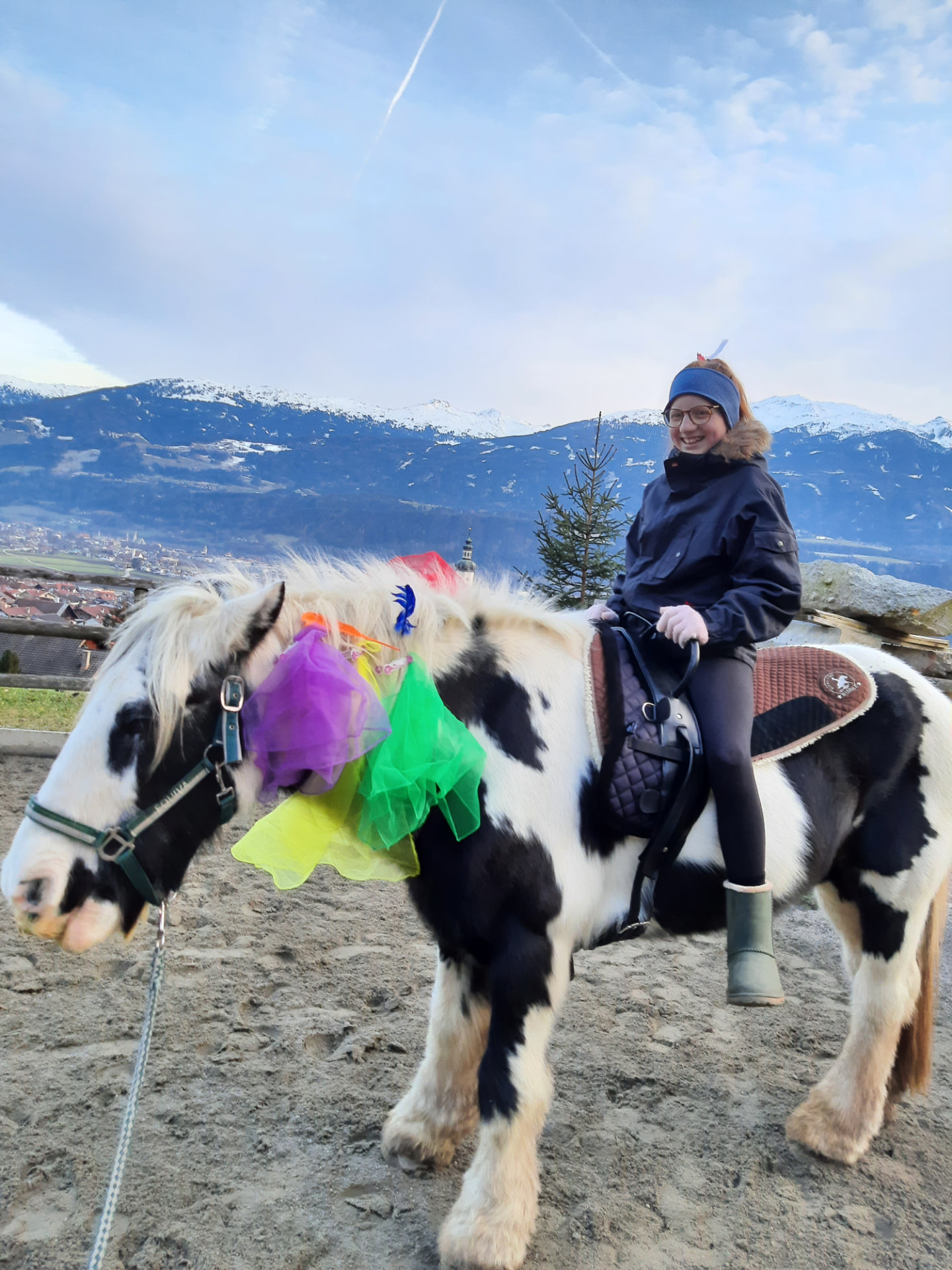 Mädchen reitet ein geschmücktes Scheckenpony mit Blick auf die winterlichen Berge